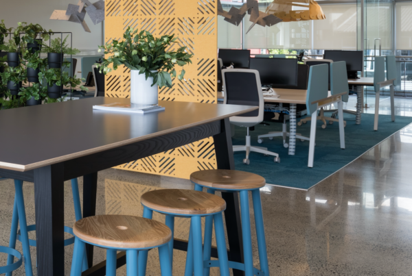 Modern open-plan office with acoustic wall paneling, shared desk spaces, and a high table with wooden stools in a collaborative workspace.