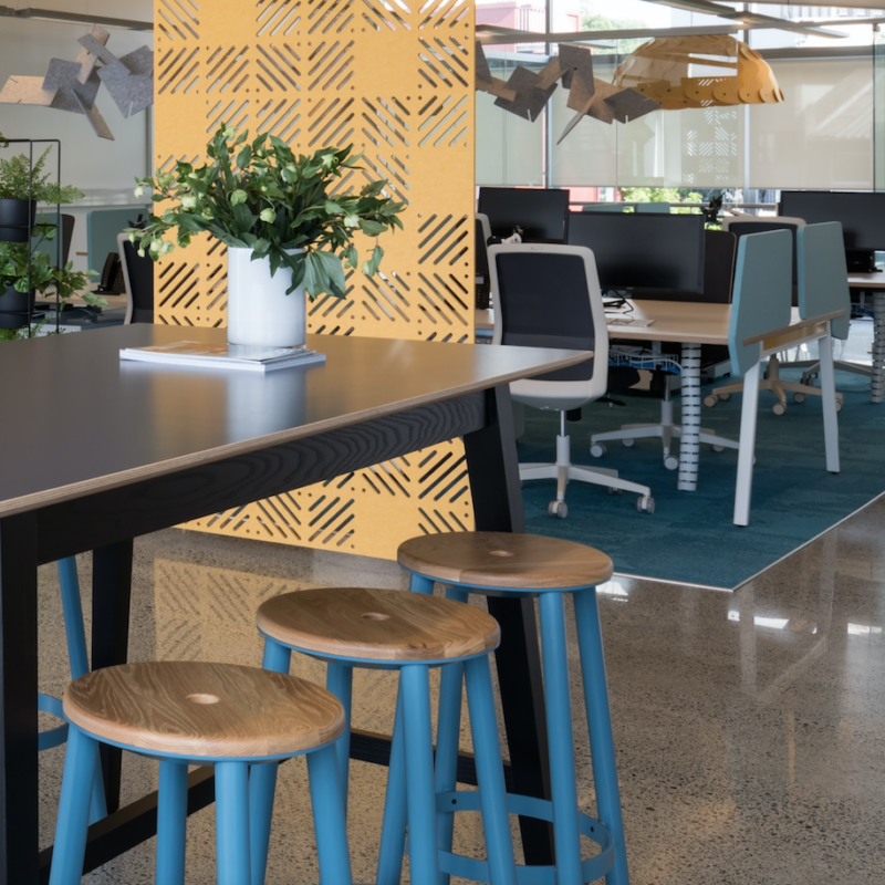 Modern open-plan office with acoustic wall paneling, shared desk spaces, and a high table with wooden stools in a collaborative workspace.