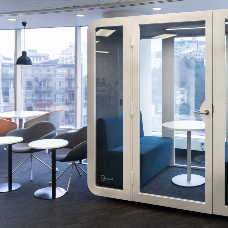 A modern office with a glass-fronted acoustic pod beside a lounge area with soft seating and city views.