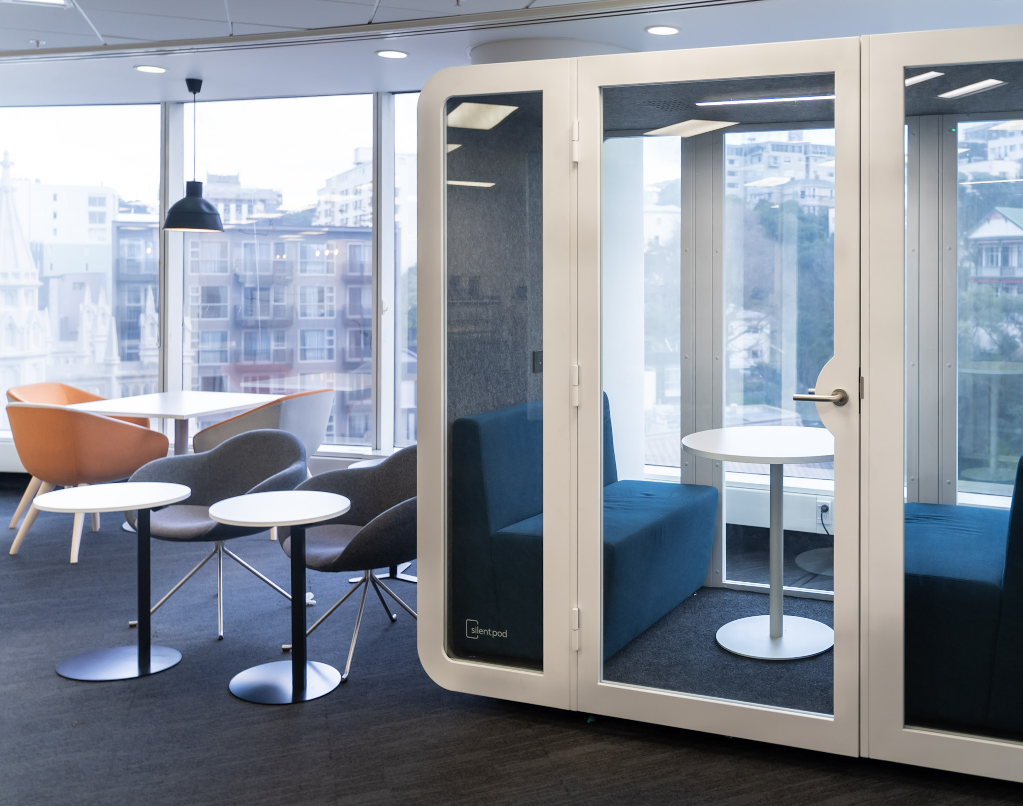 A modern office with a glass-fronted acoustic pod beside a lounge area with soft seating and city views.